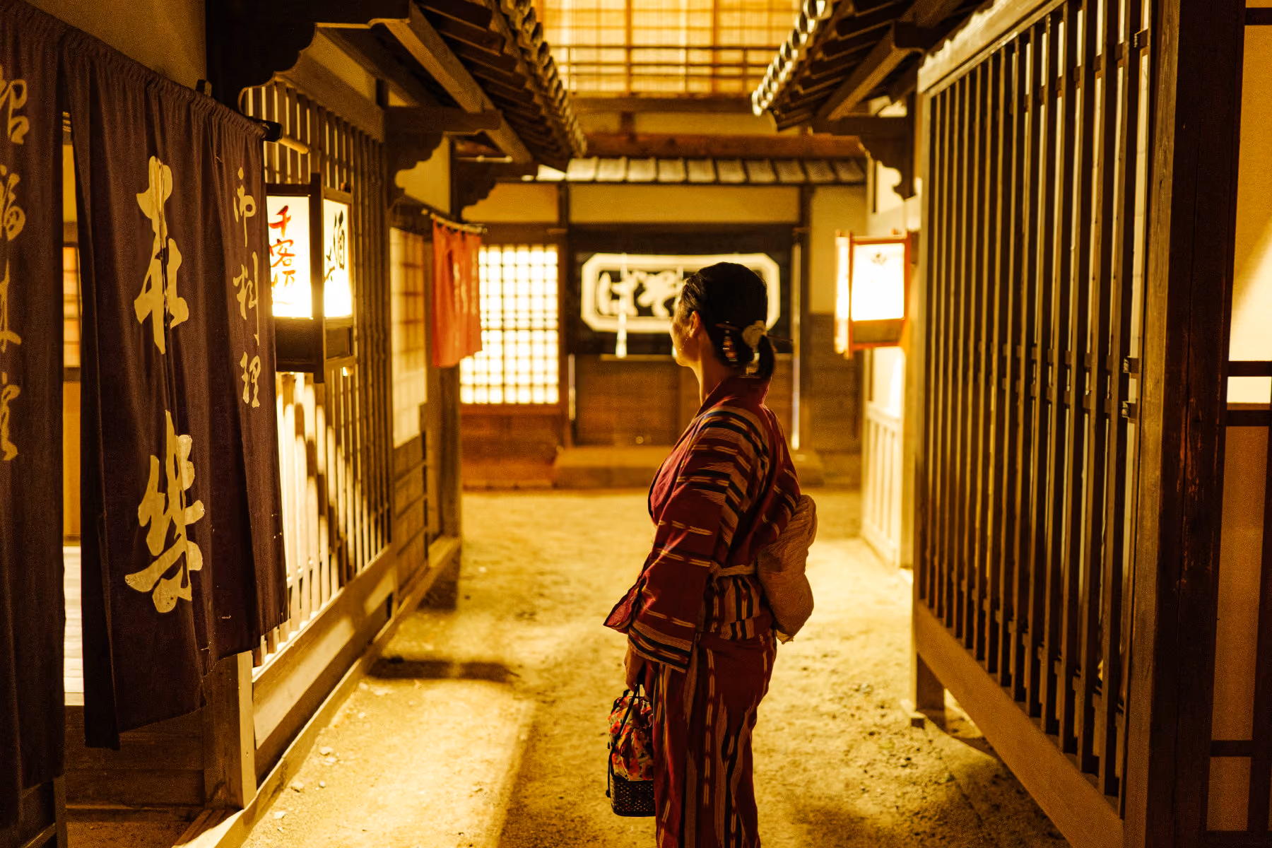 Woman in a traditional kimono standing in a warmly lit Japanese alley with wooden buildings.