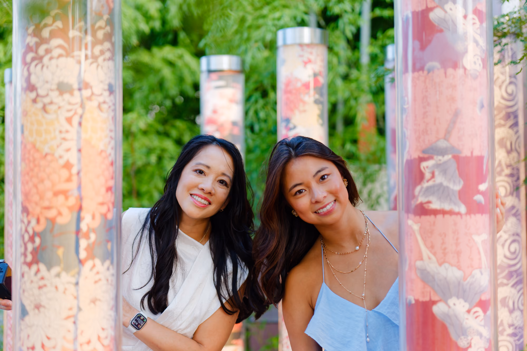 Two smiling women peek from behind decorative red and pink columns in a green outdoor setting.