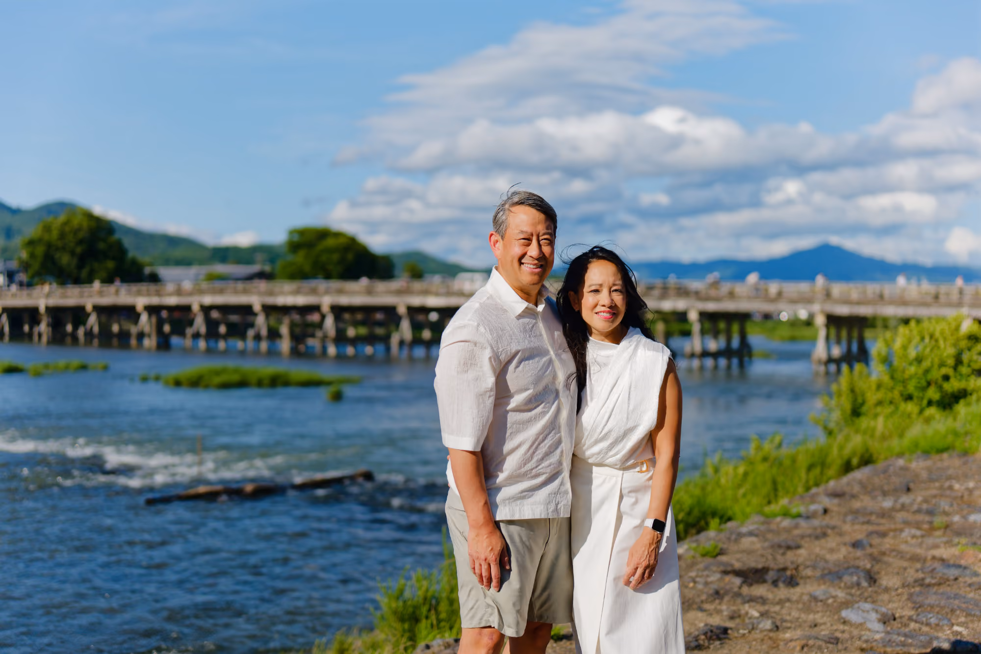 Smiling couple in white clothes posing by a river with a wooden bridge and mountains in the background.