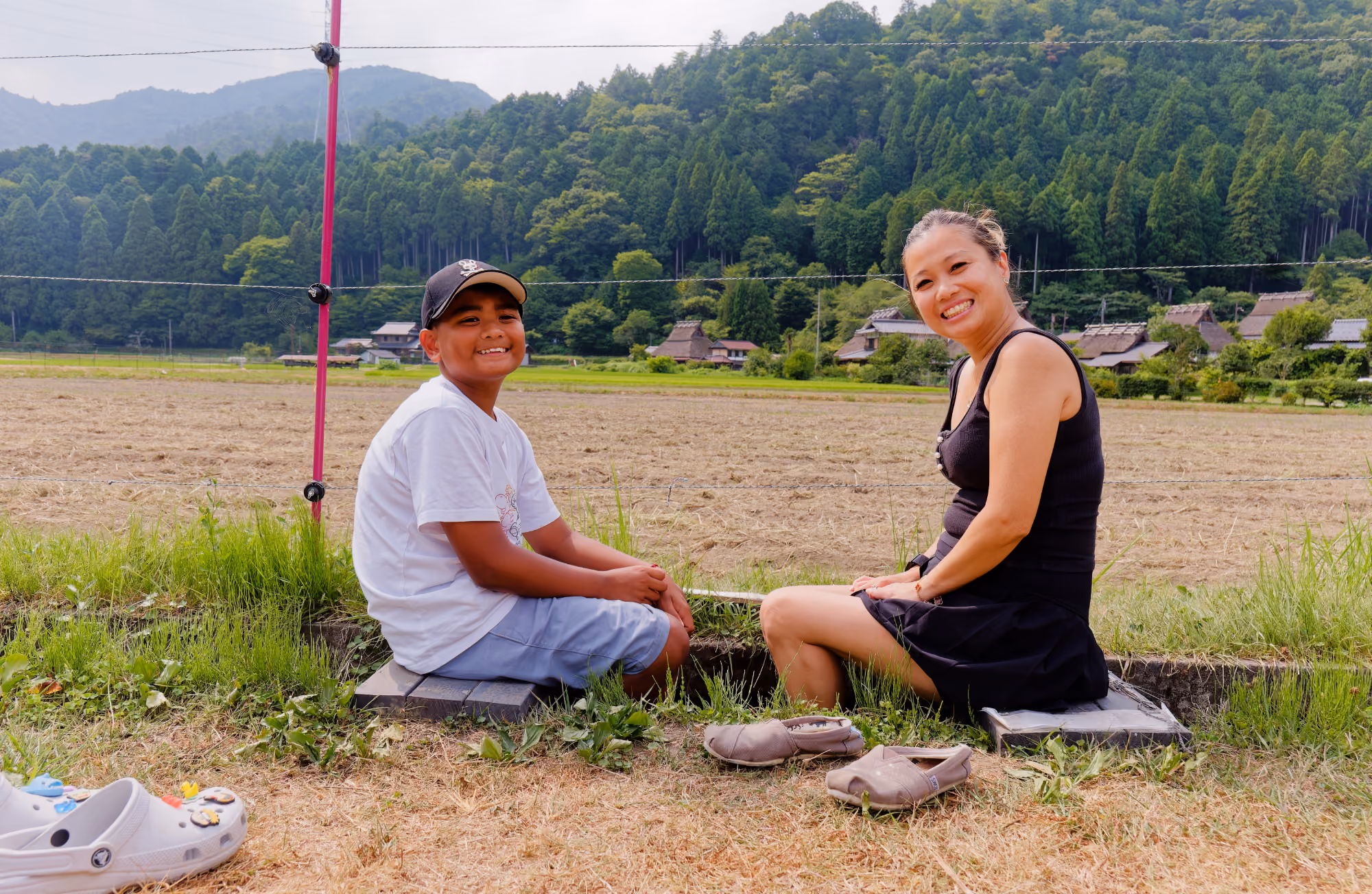 Smiling woman and boy sitting barefoot on wooden planks by a rural field with forested hills behind.