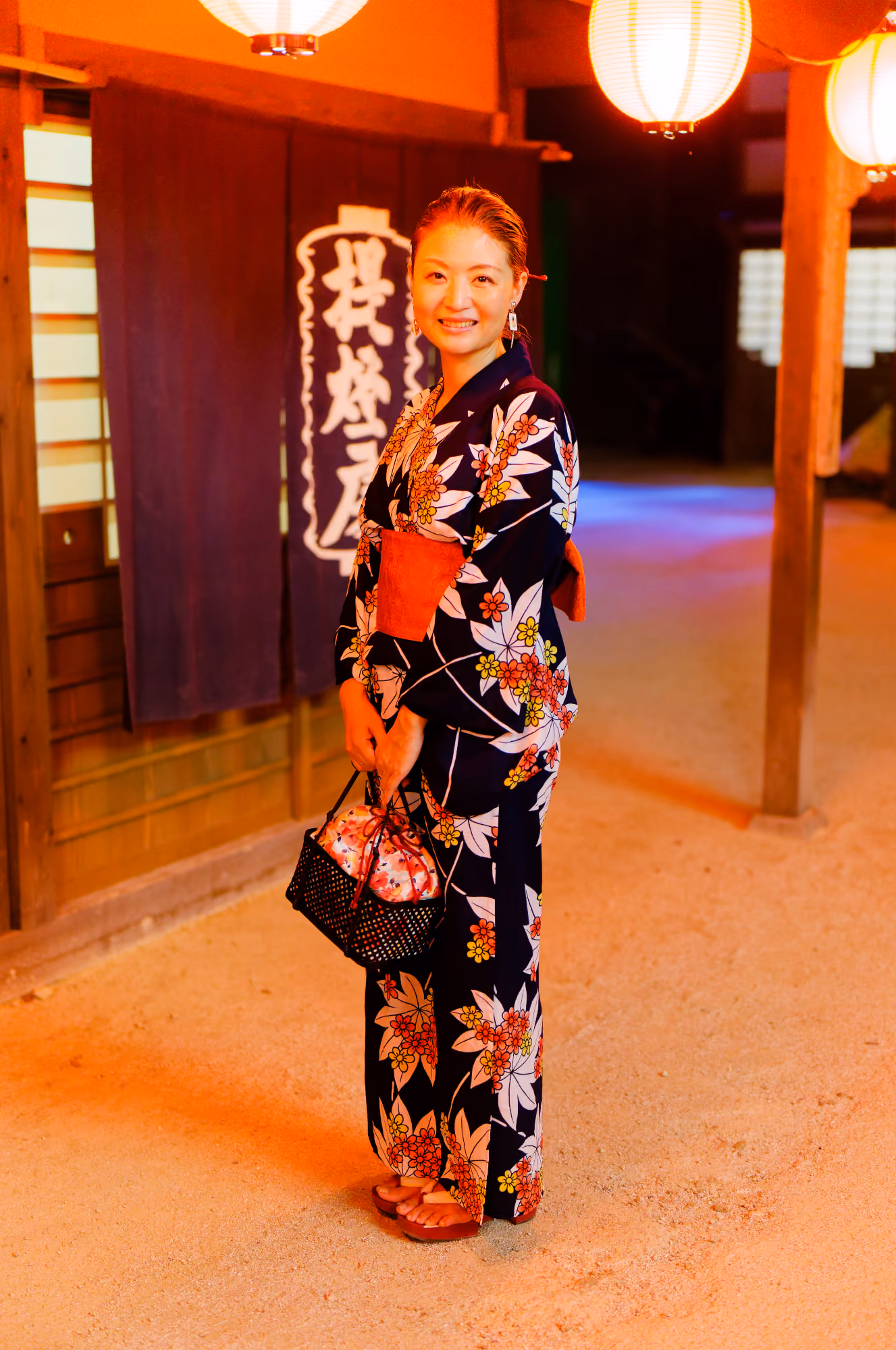 Smiling woman in a floral kimono holding a basket purse under glowing paper lanterns at night.
