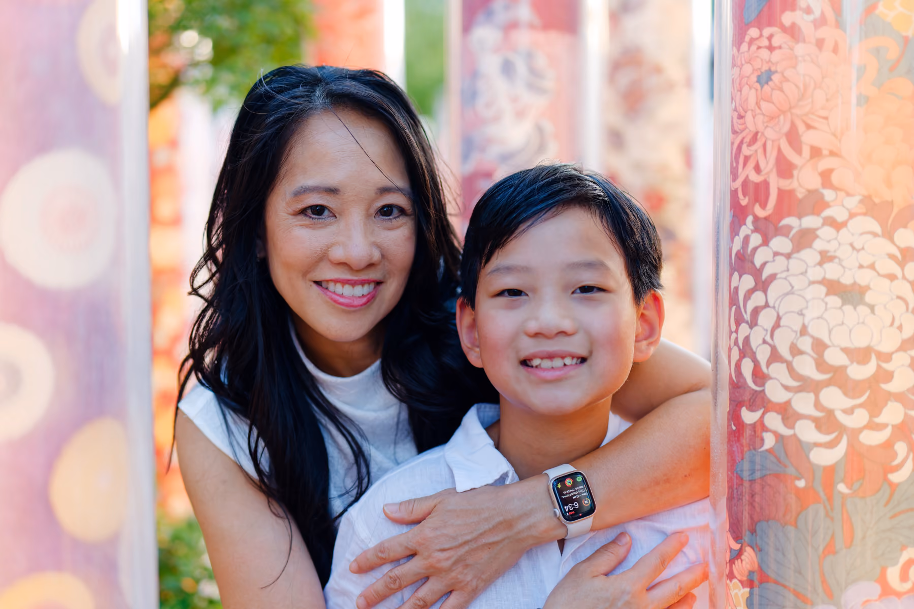 Smiling woman embraces happy boy wearing a smartwatch between colorful patterned pillars outdoors.