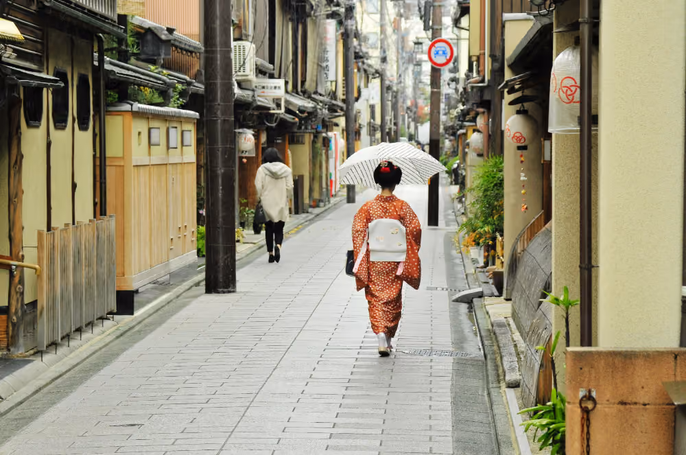 Maiko holding umbrella walking down a narrow street with another pedestrian ahead.