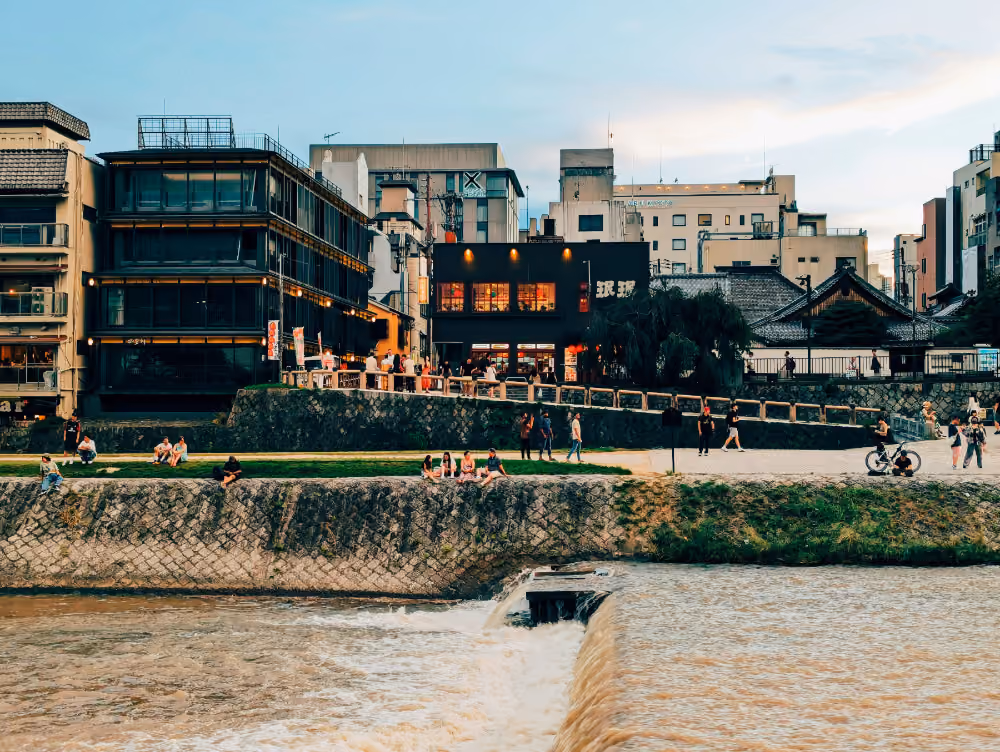 People relaxing and walking along a riverbank with urban buildings in the background at dusk.