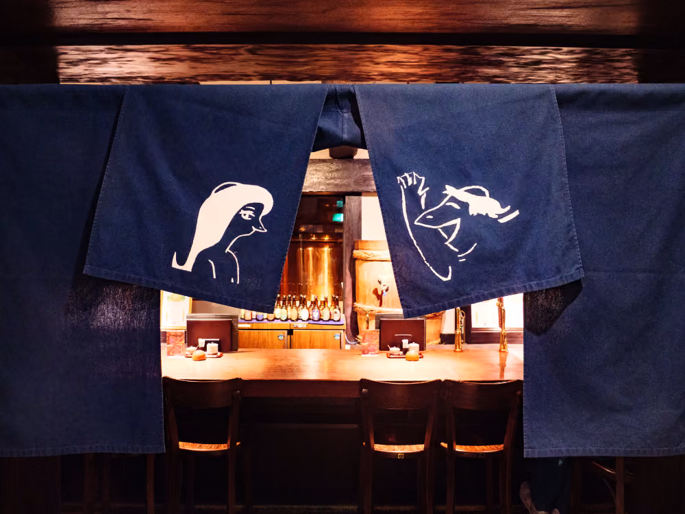 Blue fabric curtains with white abstract faces hanging at a wooden bar with stools and bottles.