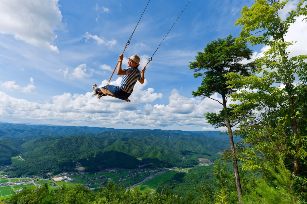 Person on a swing high above a green valley with hills under a partly cloudy blue sky.