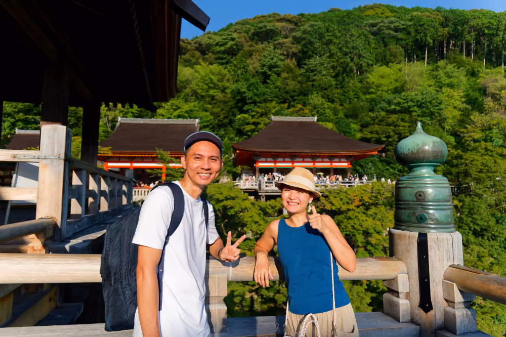 Two tourists on a wooden balcony overlooking a crowded traditional Japanese temple in lush greenery.