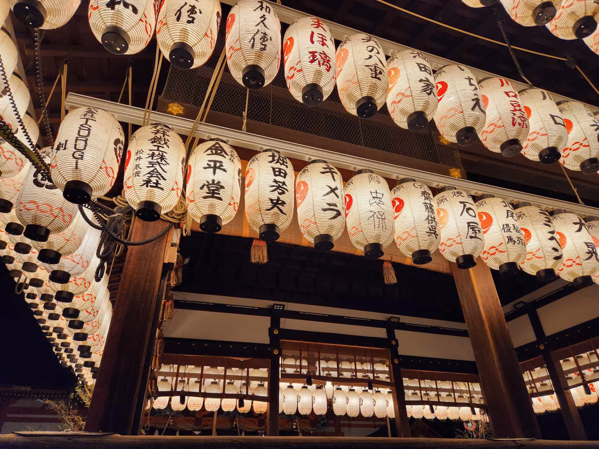 Rows of illuminated Japanese paper lanterns with black and red kanji hanging under wooden beams at night.
