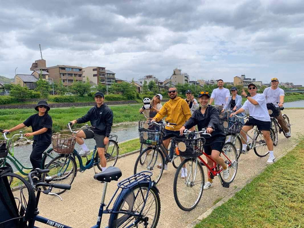 Group of people smiling on bicycles on a riverside path with buildings and cloudy sky in background.