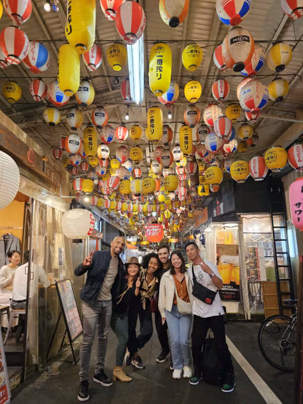 Group of six friends posing and smiling under hanging red, yellow, and white lanterns at night.