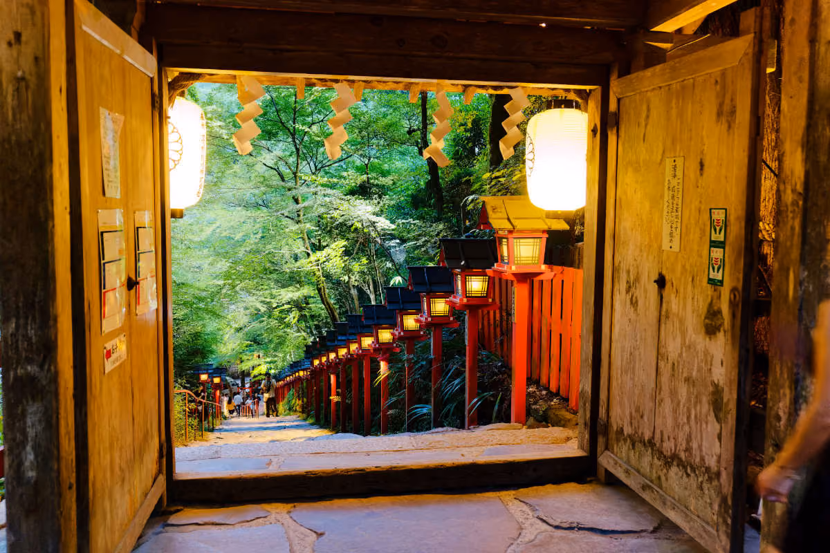 View through wooden gate to stone steps lined with red lanterns and green trees in a serene outdoor setting.