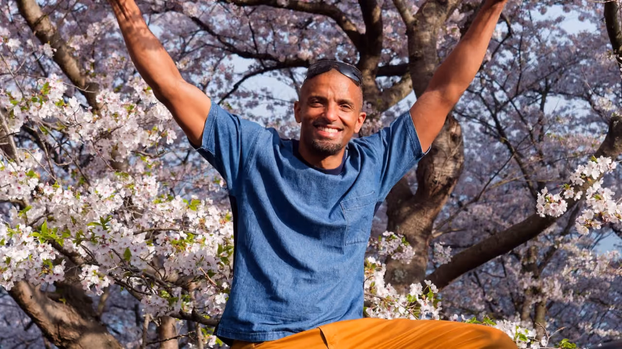 Smiling man in blue shirt with arms raised sitting among blooming cherry blossom trees.