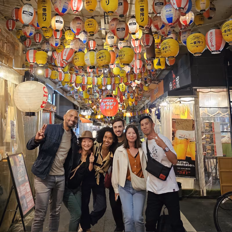 Group of six friends posing and smiling under hanging red, yellow, and white lanterns at night.