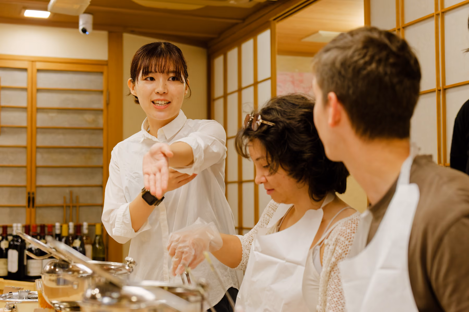 A woman in a white shirt is explaining or demonstrating something with her arm extended to two seated people wearing aprons in a traditional Japanese room with sliding wooden doors and bottles in the background.