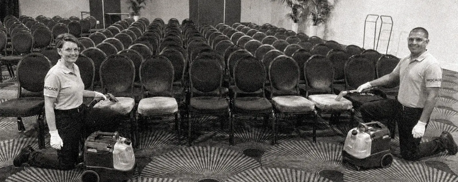 Deux techniciens Mocade en polo clair nettoient à genoux les chaises d'une salle de réception, photographie d'archive en noir et blanc