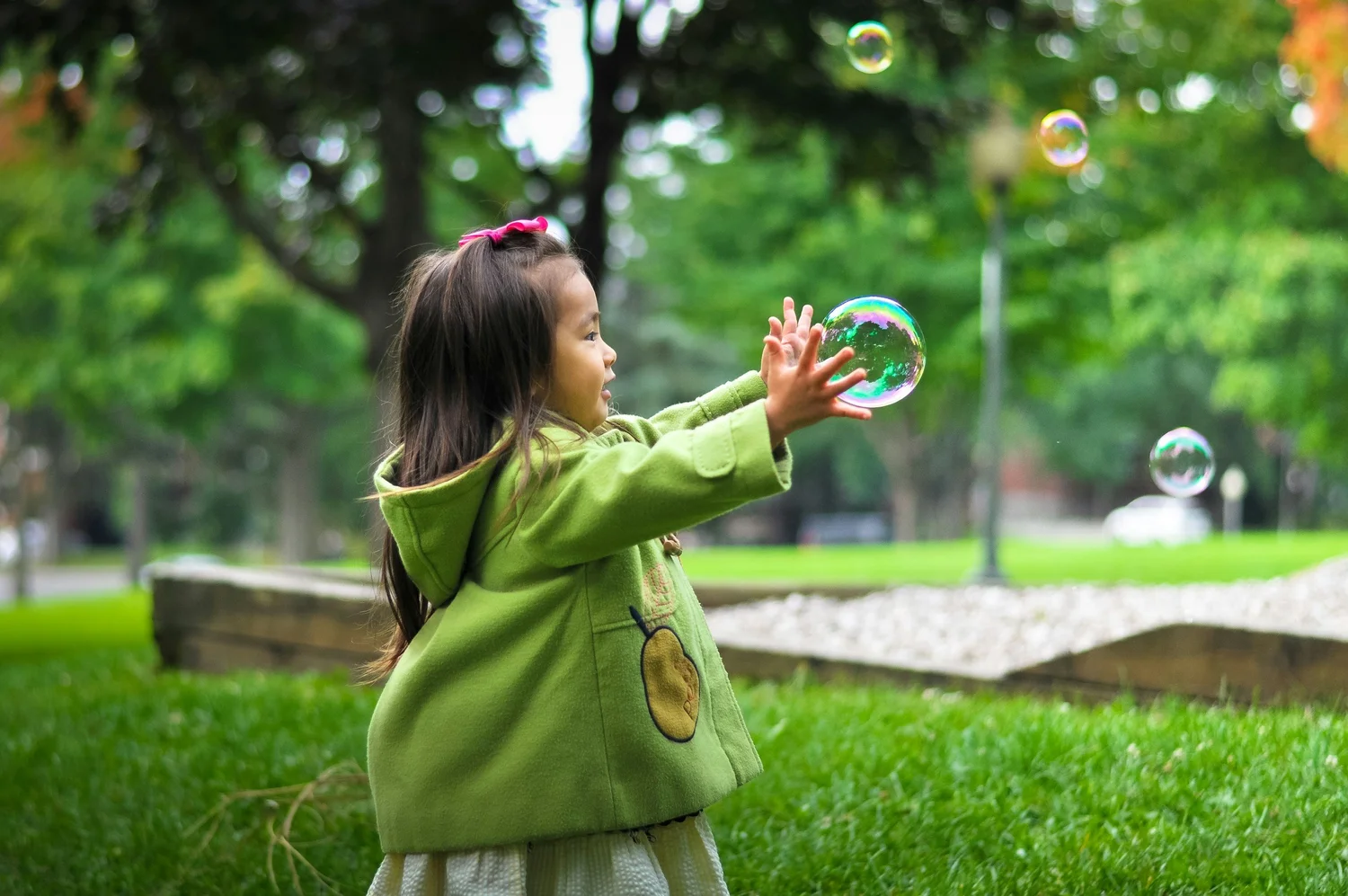 A young child in a green coat reaching for soap bubbles in a park