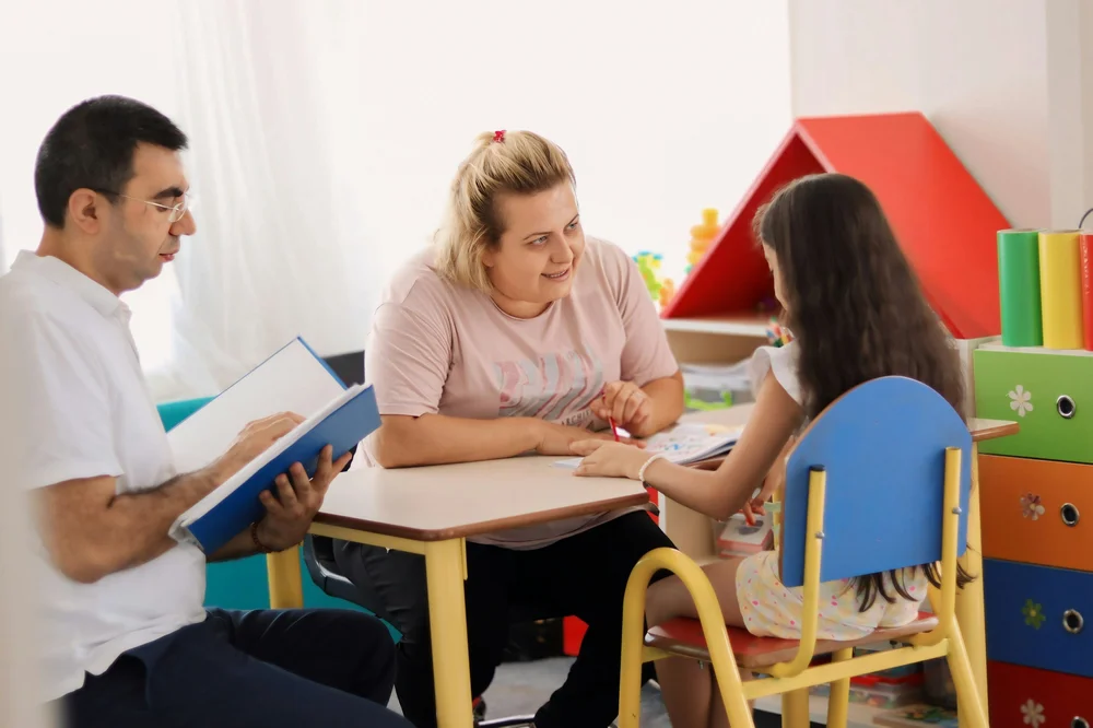 Two adults and a child sitting at a small table in a playroom looking at a book