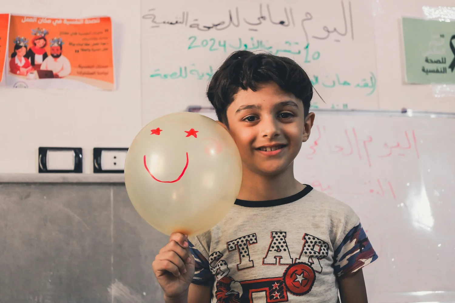 A boy in a classroom holding up a balloon with a smiling face drawn on it