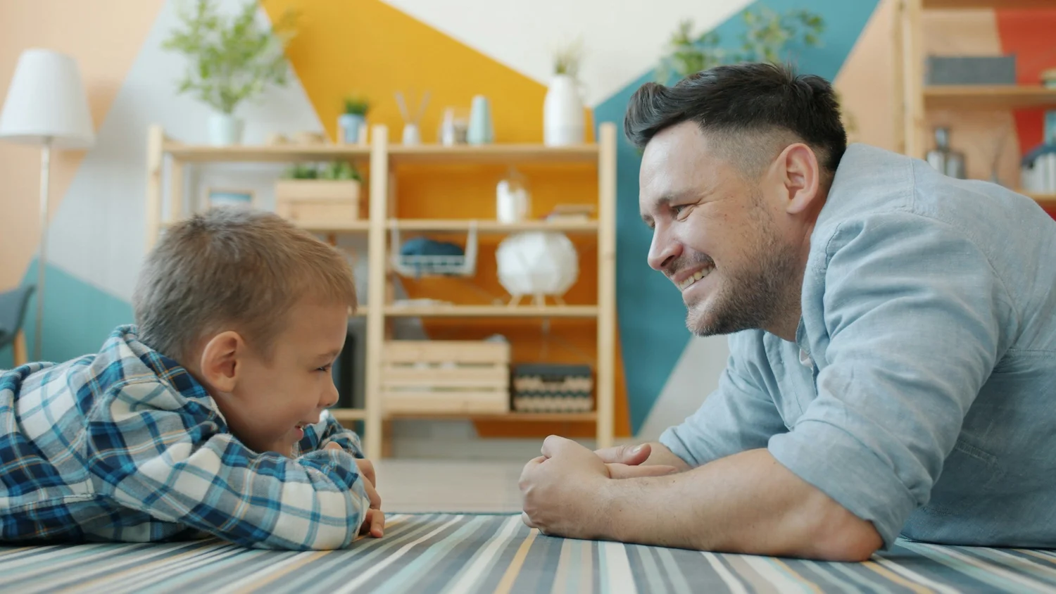 An adult and a child lying face to face on the floor of a playroom