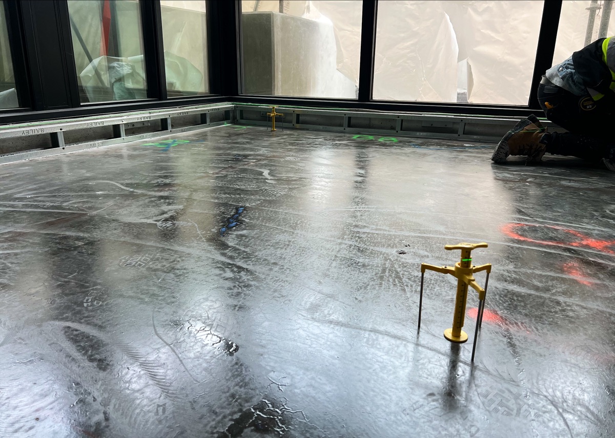 Worker kneeling at the edge of a wet levelling compound floor, yellow gauge in the foreground