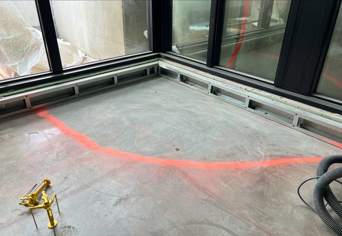 Grey subfloor with an orange spray line near the window wall, gauges and a vacuum hose on site