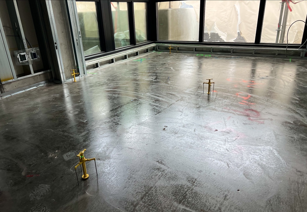 Freshly poured levelling compound across a corner room with floor-to-ceiling windows, yellow gauges standing on the floor; illustrative point markers overlaid