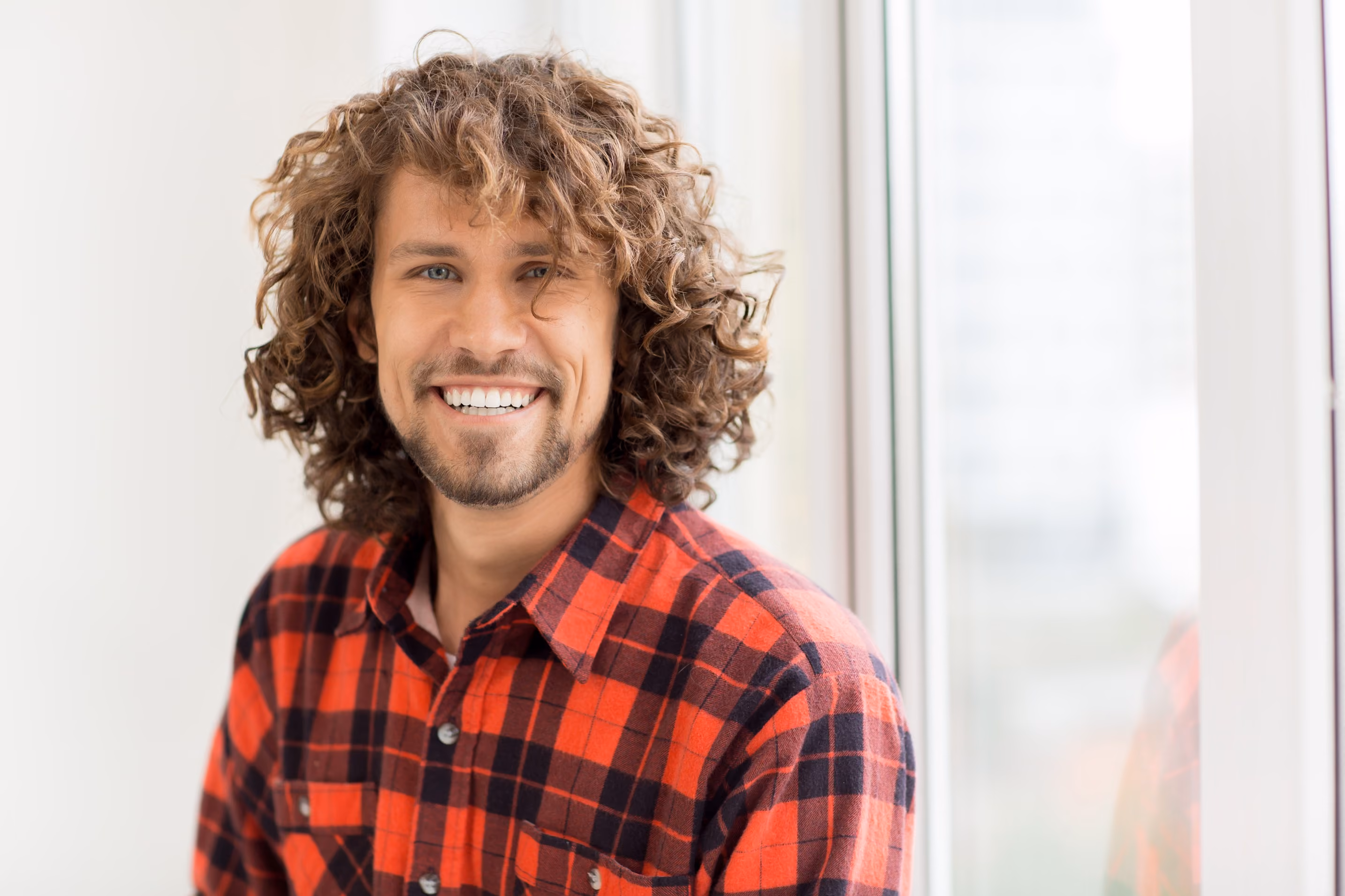Smiling man with curly brown hair wearing a red and black plaid shirt, standing indoors near a bright window.