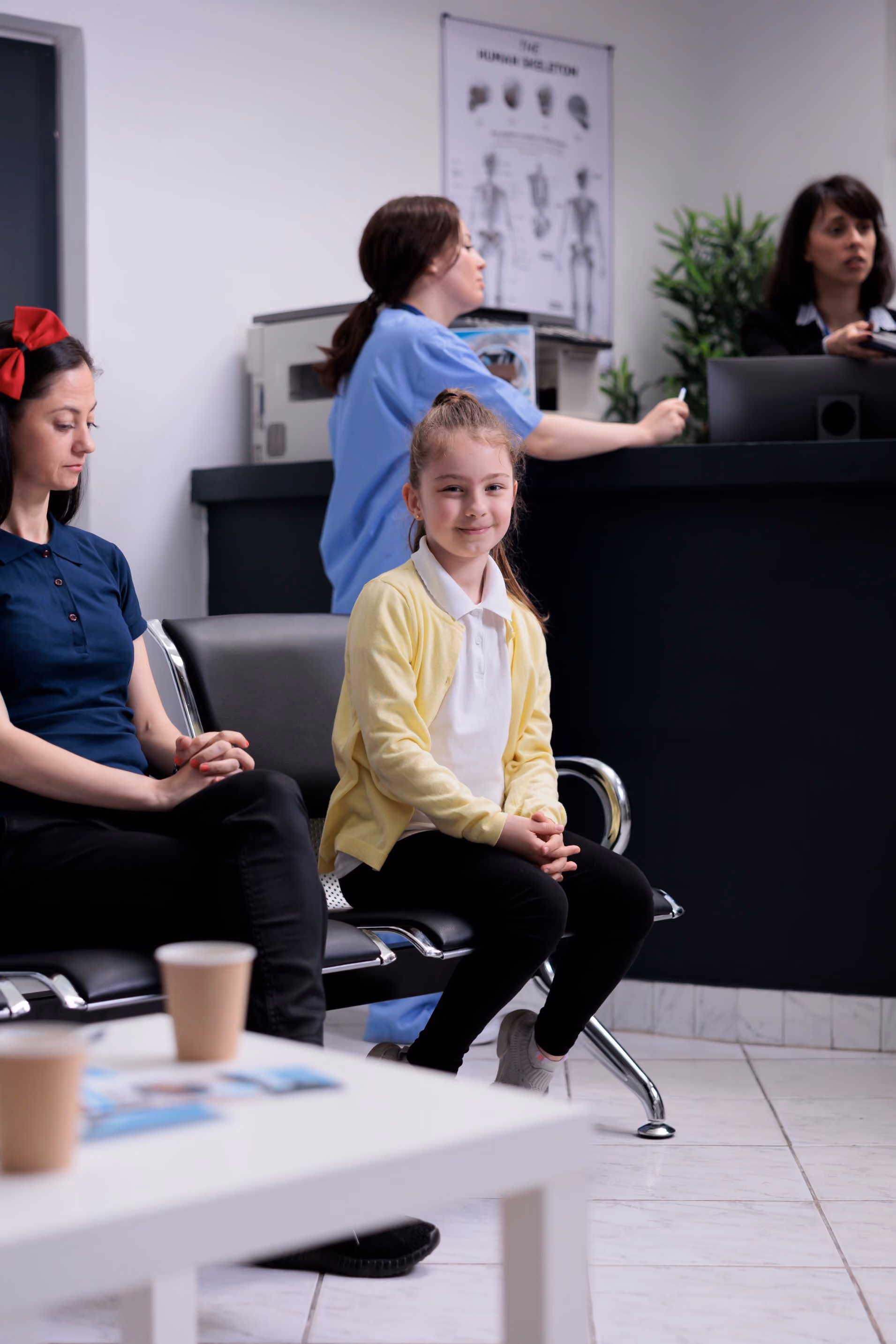 Young girl in a yellow cardigan sitting beside an adult in the waiting room of a dental clinic while staff assist patients at the reception desk.