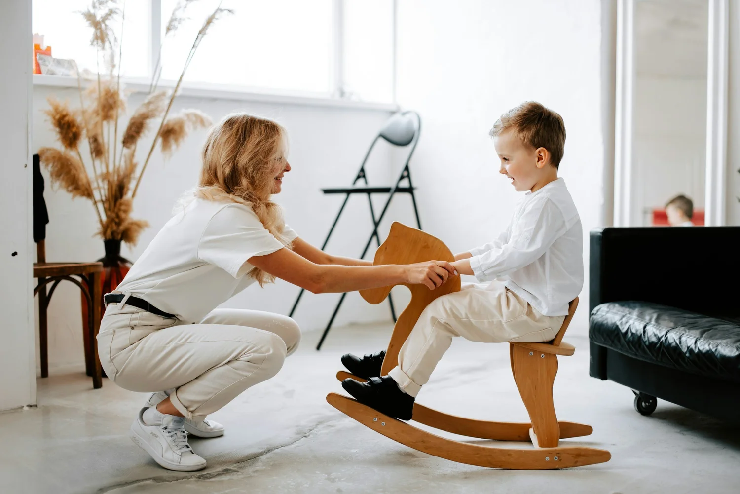 An adult kneeling beside a young child on a rocking horse in a living room