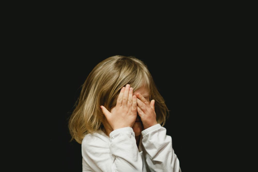 A child standing against a dark background with both hands over their face