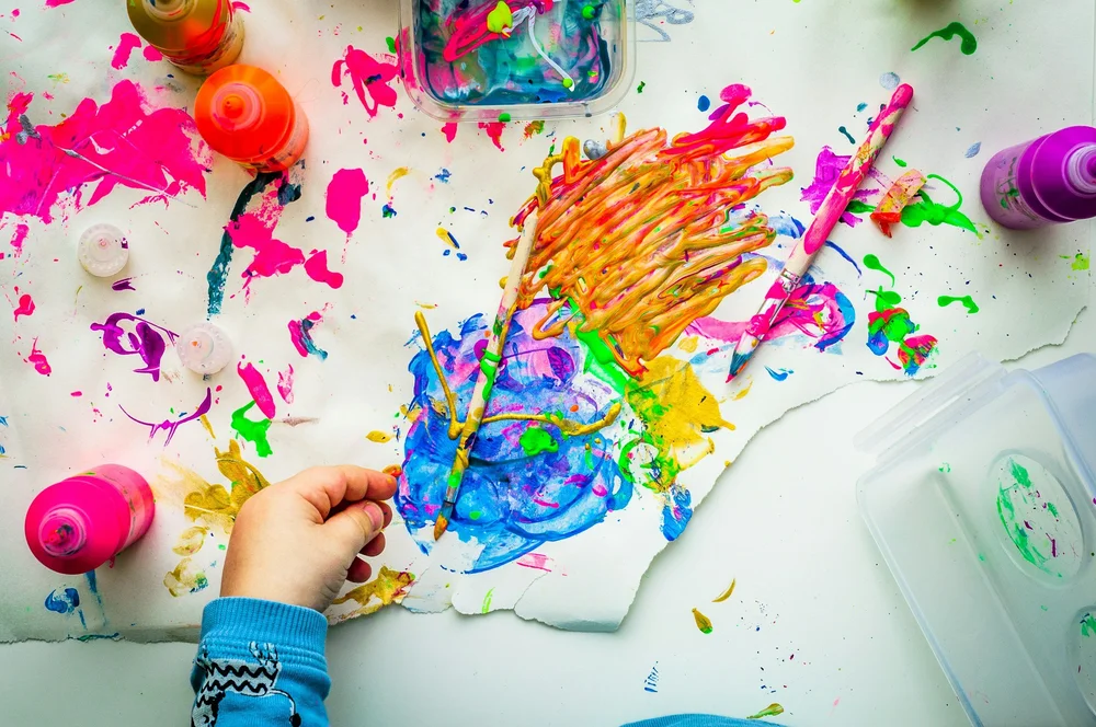 A child's hand painting bright swirls on paper surrounded by paint pots