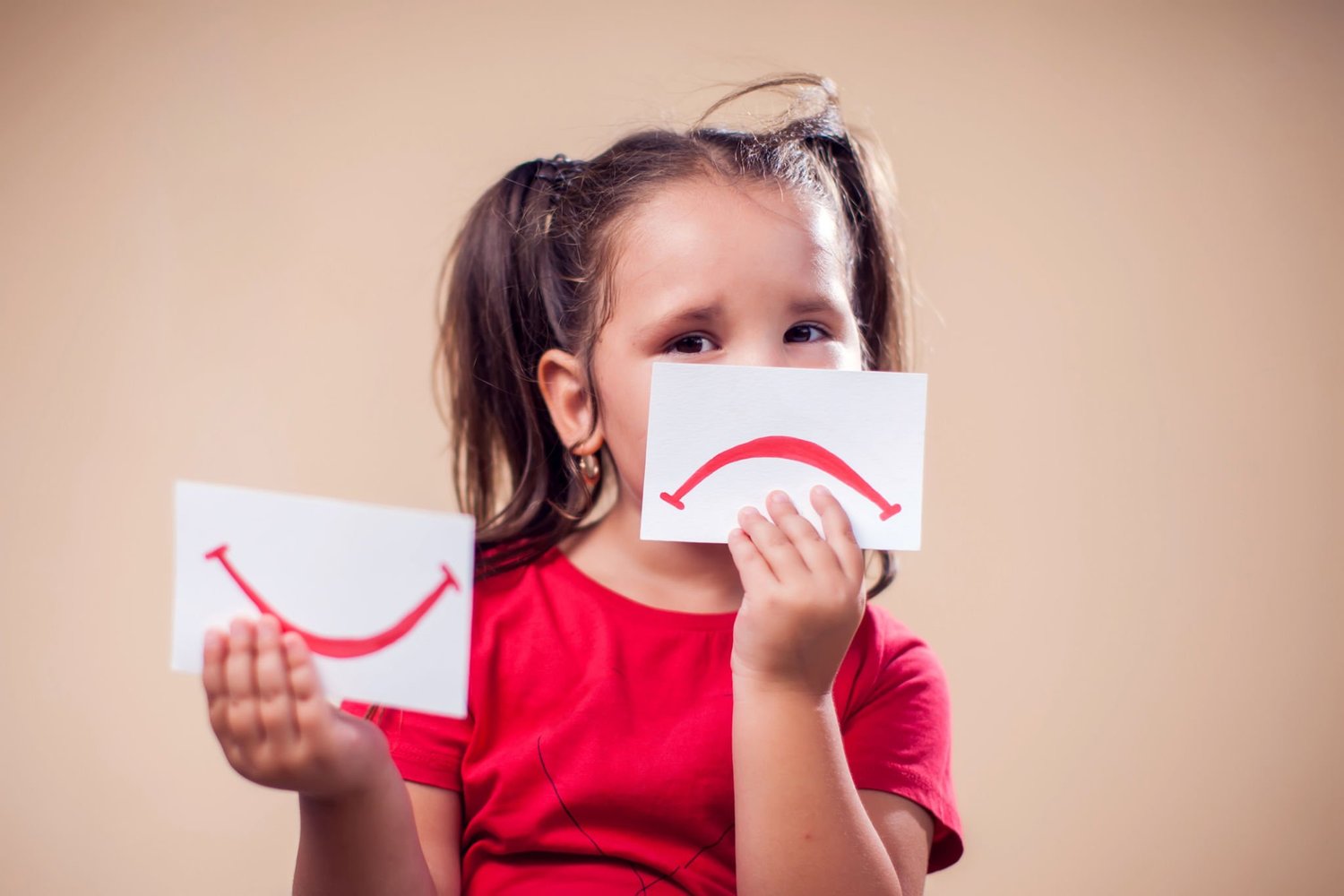 A child holding a paper smile and a paper frown up to their face