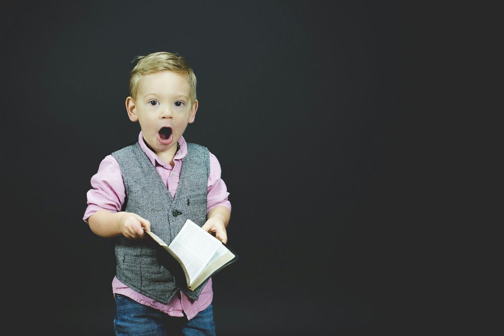 A boy in a waistcoat holding an open book with a surprised expression
