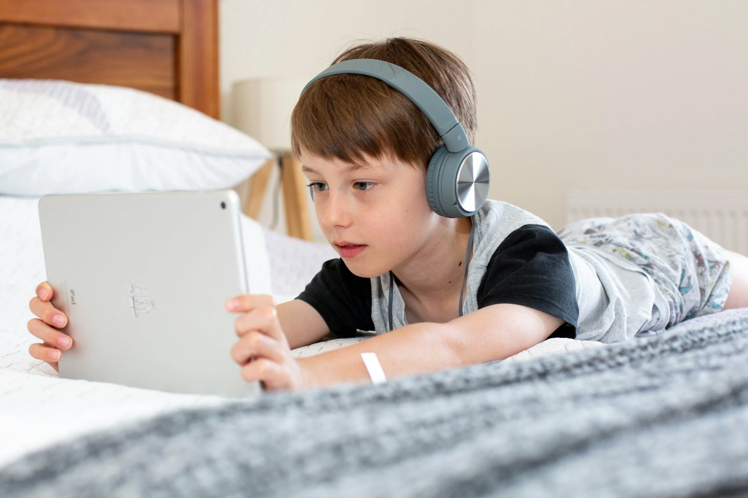 A boy lying on a bed wearing headphones and watching a tablet