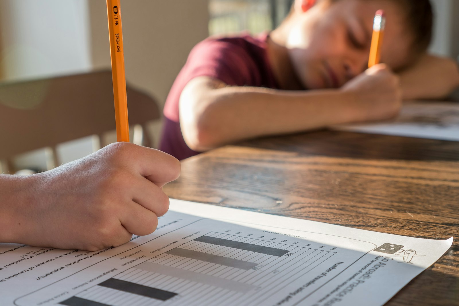 Two children at a wooden table with pencils and worksheets, one slumped over, homework meltdowns