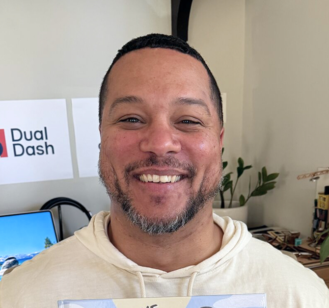 Smiling man with short dark hair and a trimmed beard wearing a light-colored hoodie, standing indoors in front of a wall with a Dual Dash sign and a desk with plants and books.