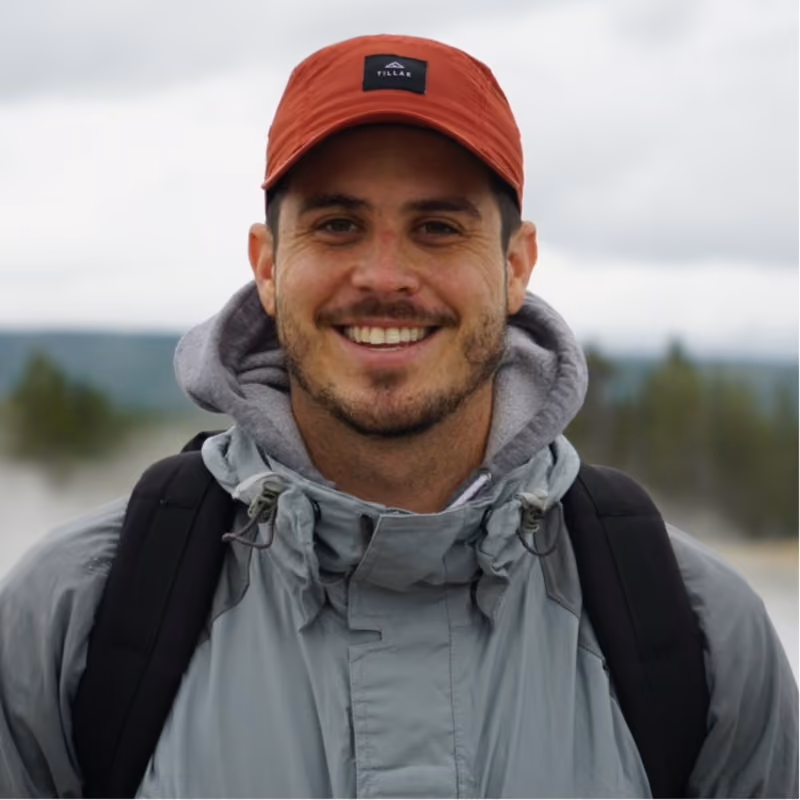 Smiling man outdoors wearing a red cap, gray jacket with a hood, and a black backpack with a blurred natural background.