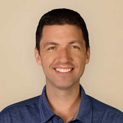 Smiling man with short dark hair wearing a blue collared shirt against a light beige background.