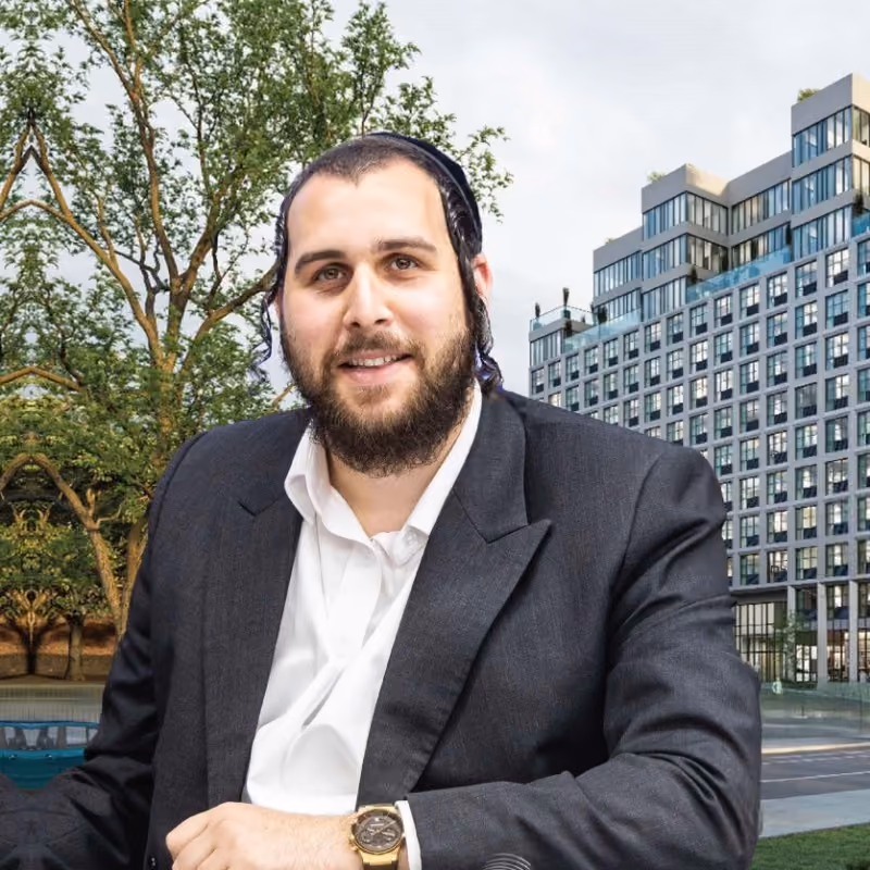 Smiling man with dark hair, beard, and sidelocks wearing a black blazer, white shirt, and gold watch seated outdoors with trees and a modern multi-story building in the background.