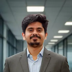 Smiling man with dark hair and beard wearing a gray blazer and light blue shirt, standing in a bright corridor with ceiling lights and glass walls.
