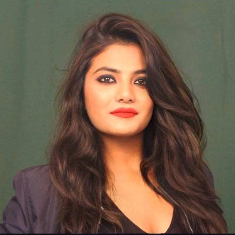 Portrait of a woman with long wavy dark hair, wearing a black top and jacket, with red lipstick and dark eye makeup, against a plain green background.