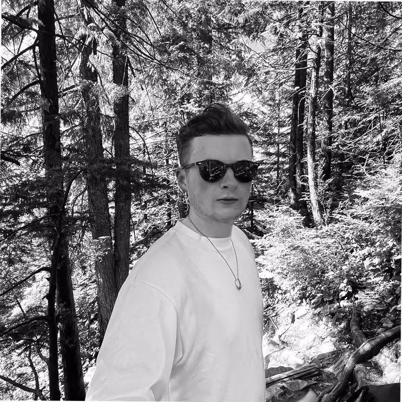 Young man wearing sunglasses and a light-colored long-sleeve shirt standing in a forest with tall trees and sunlight filtering through the leaves.