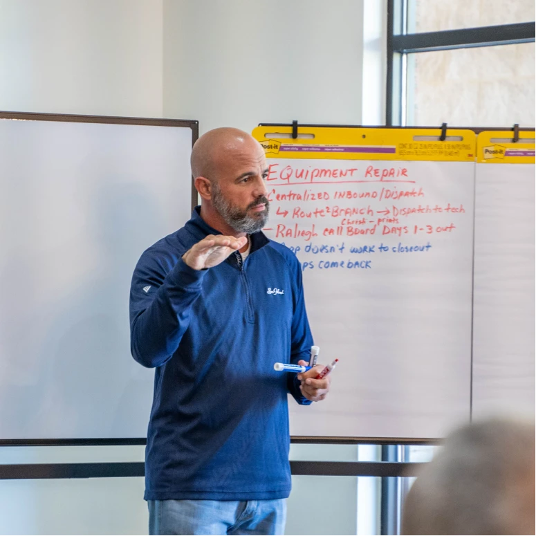 Man with a beard and shaved head wearing a navy blue pullover stands in front of a whiteboard with handwritten notes about equipment repair while holding three markers and gestures with his other hand as if explaining.