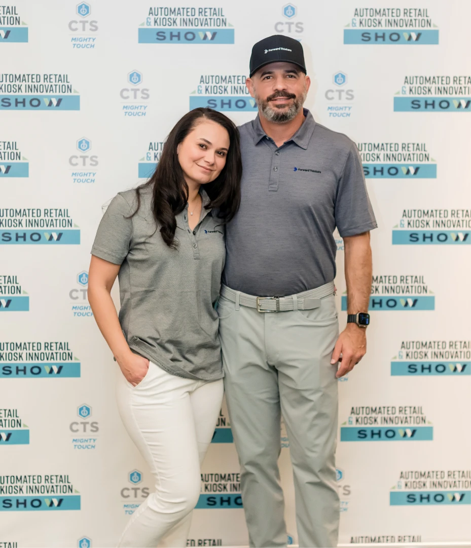 A smiling woman and man stand close together in front of a backdrop with repeated text 'Automated Retail & Kiosk Innovation Show' and logos, both wearing gray company polo shirts; the man also wears a matching cap and a smartwatch.