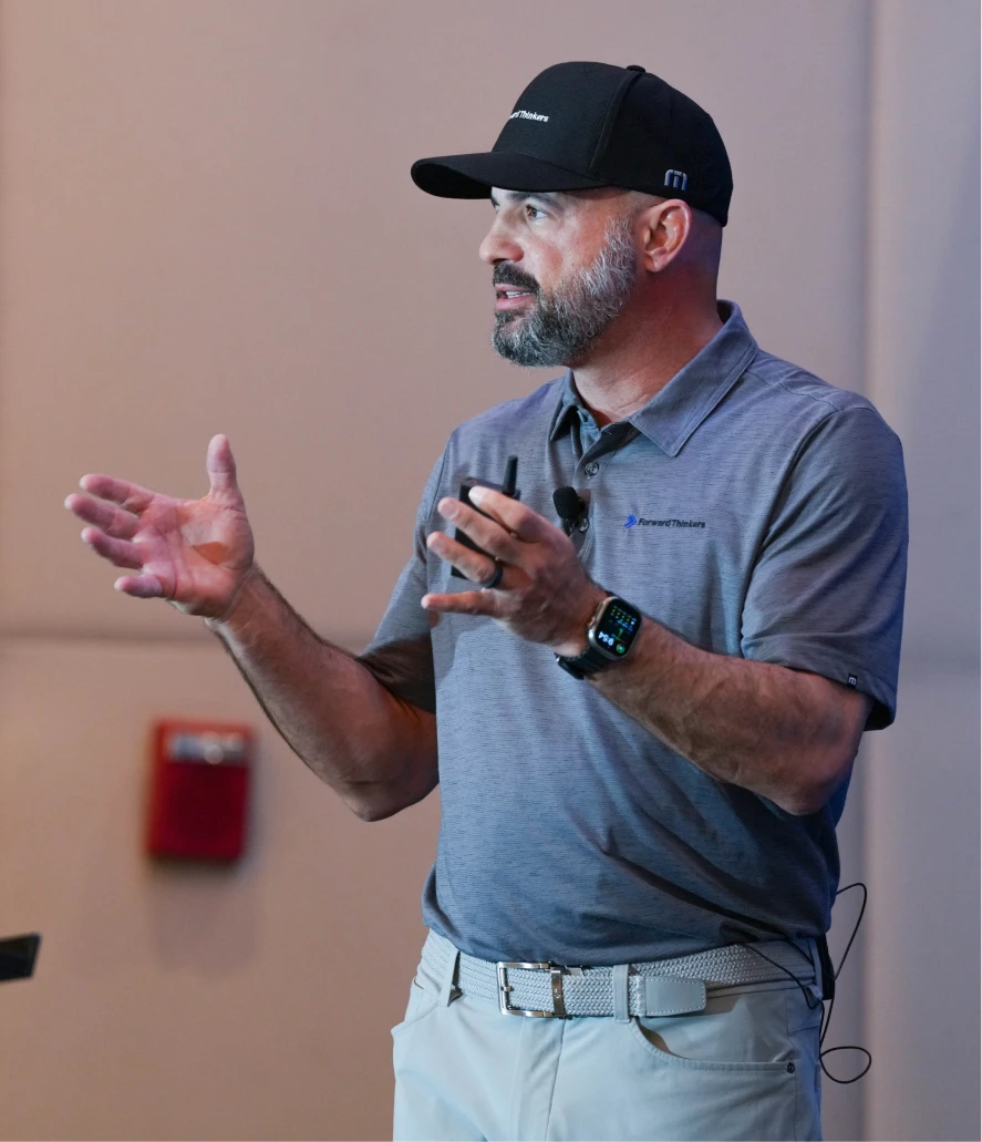 A man with a beard wearing a black cap, gray polo shirt with Forward Thinkers logo, light pants, and a smartwatch speaks and gestures with his hands indoors.