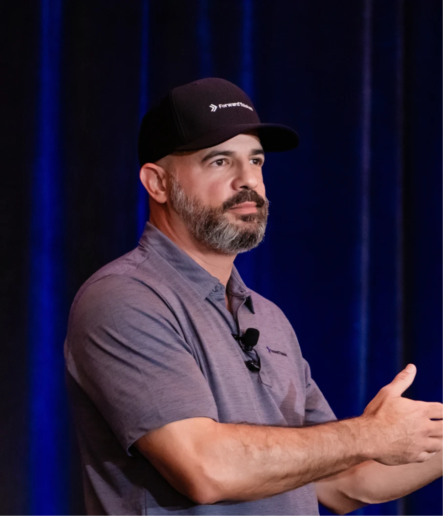 Man with a beard wearing a black cap and a gray polo shirt speaking in front of a dark blue curtain backdrop.