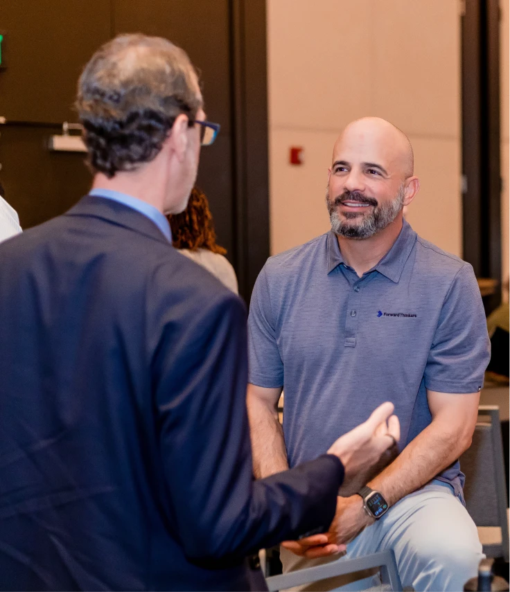 Two men engaged in conversation indoors, one in a blue polo shirt with a beard smiling, the other in a suit with glasses seen from the back.