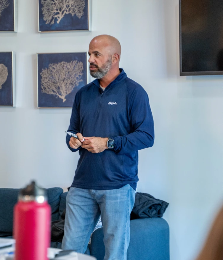 A bald man with a beard stands wearing a navy blue pullover and light blue jeans, holding a marker in his hands in a room with framed coral artwork on the wall and a television mounted on the right.