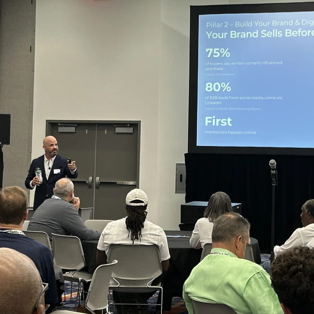 A man in a dark blazer holding a bottled drink and a pointer is giving a presentation to an audience seated in a conference room, with a projection screen displaying a slide titled 'Your Brand Sells Before' showing statistics about buyer influence and social media leads.