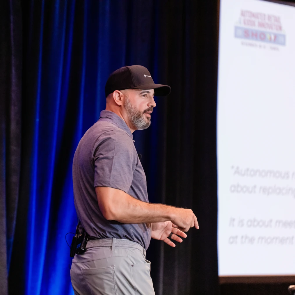 A man wearing a gray polo shirt, light gray pants, and a black cap is speaking on stage with a microphone attached to his back belt, standing in front of a presentation screen and dark blue curtains.